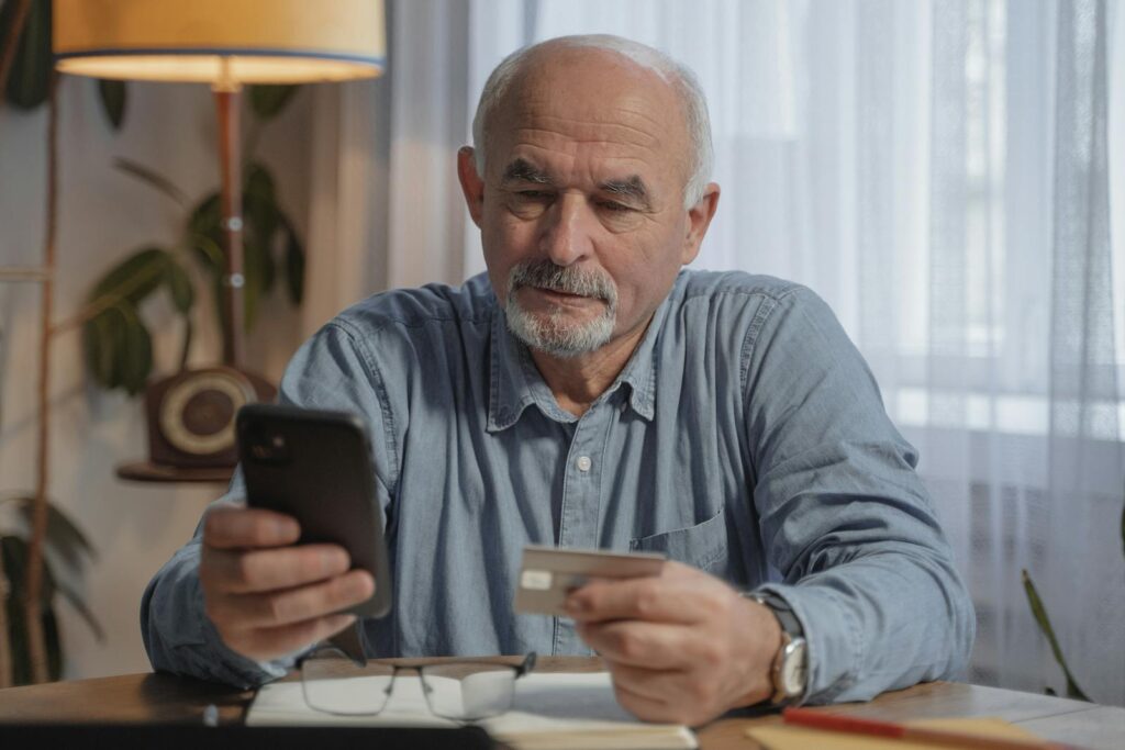 An elderly man using a smartphone and credit card for online shopping at home.